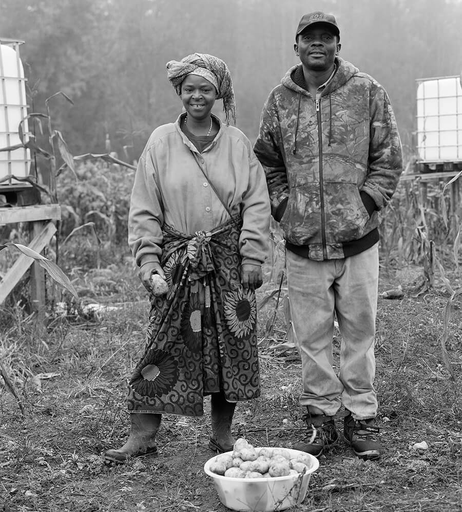 black and white photo of a male and female refugee standing and smiling on a farm with a basket of potatoes